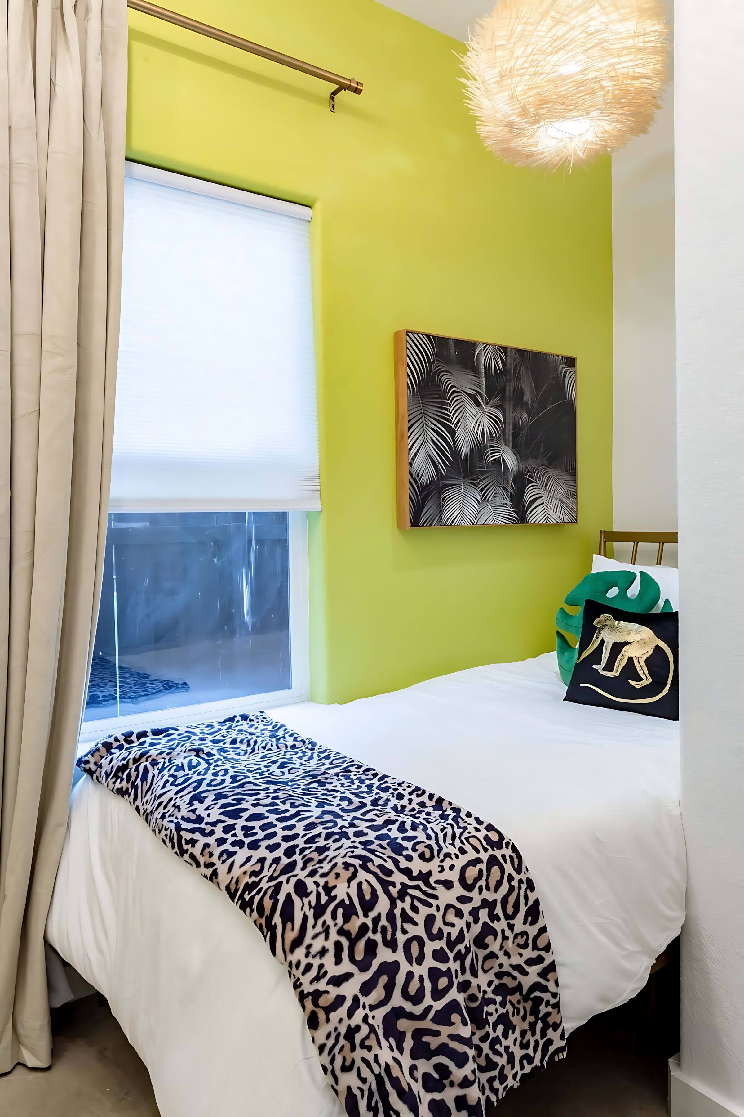 A bedroom with a bed, a window with curtains, a wall painting, and a hanging light fixture.