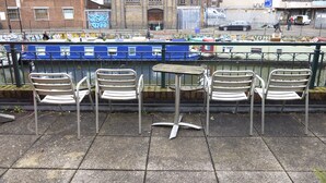 A row of metal chairs and a small table on a paved area with a metal railing in the background.