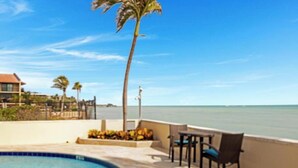 Zona de piscina con vista al océano, una palmera y una mesa con sillas.