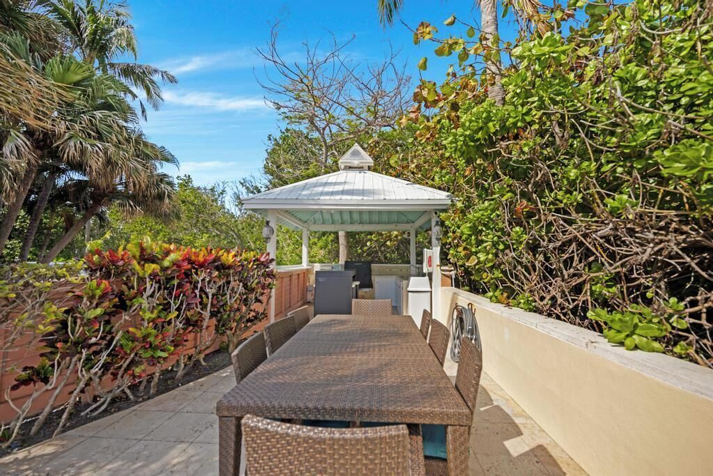 A covered outdoor seating area with a table and chairs, surrounded by lush greenery and a gazebo structure.