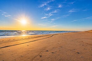 Una playa con huellas que conducen al agua, cielo despejado y el sol poniéndose o saliendo.