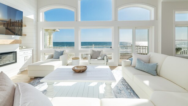 A spacious living room with a large white sofa, a coffee table, and a fireplace. There are multiple windows offering a view of the ocean. - 🌊 Ultra-Luxury OCEANFRONT | Private Heated Pool🏖️Hot Tub & Unmatched Views! (Corolla)