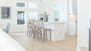 A modern kitchen with white cabinetry, a central island with bar stools, and large windows providing natural light. - 🌊 Ultra-Luxury OCEANFRONT | Private Heated Pool🏖️Hot Tub & Unmatched Views! (Corolla)