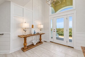 A spacious room with a wooden console table, white walls, and a large glass door leading to an outdoor area. - 🌊 Salty Treat | Luxury Oceanfront | Private Heated Pool⛱️Hot Tub & Ocean Views (Corolla)