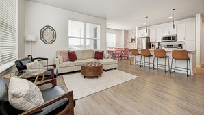 Ein modernes Wohnzimmer mit einer Couch, einem Hocker und Sesseln, eine Küche mit Barhocker und großen Fenstern.