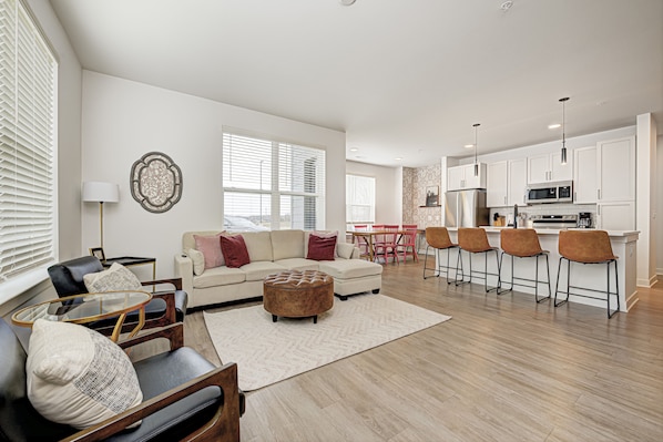 A modern living room with a sofa, ottoman, and chairs, a kitchen with bar stools, and large windows.