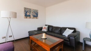 A modern living room with a black leather sofa, a glass coffee table, and two wall-mounted paintings. - Upper Views 2D Wifi, Balcony & Parking (Las Condes)