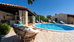 Une piscine en pierre, des chaises longues, un fauteuil en osier et un bâtiment en pierre surmonté d’un toit en tuiles.