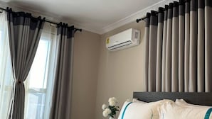 A hotel room with a bed, a window with curtains, an air conditioning unit, and a bedside table with a vase of flowers.