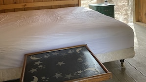 A wooden cabin room with a bed, a small table with a star and moon design, and a window with a forest view. - HARVEST MOON - ROMANTIC - OFF GRID - WINE TRAIL CABIN - Pet Friendly! 4 Seasons (Prattsburgh)