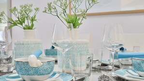 Una mesa dispuesta con vajilla azul y blanca estampada, copas de cristal transparente y pequeñas macetas con plantas.