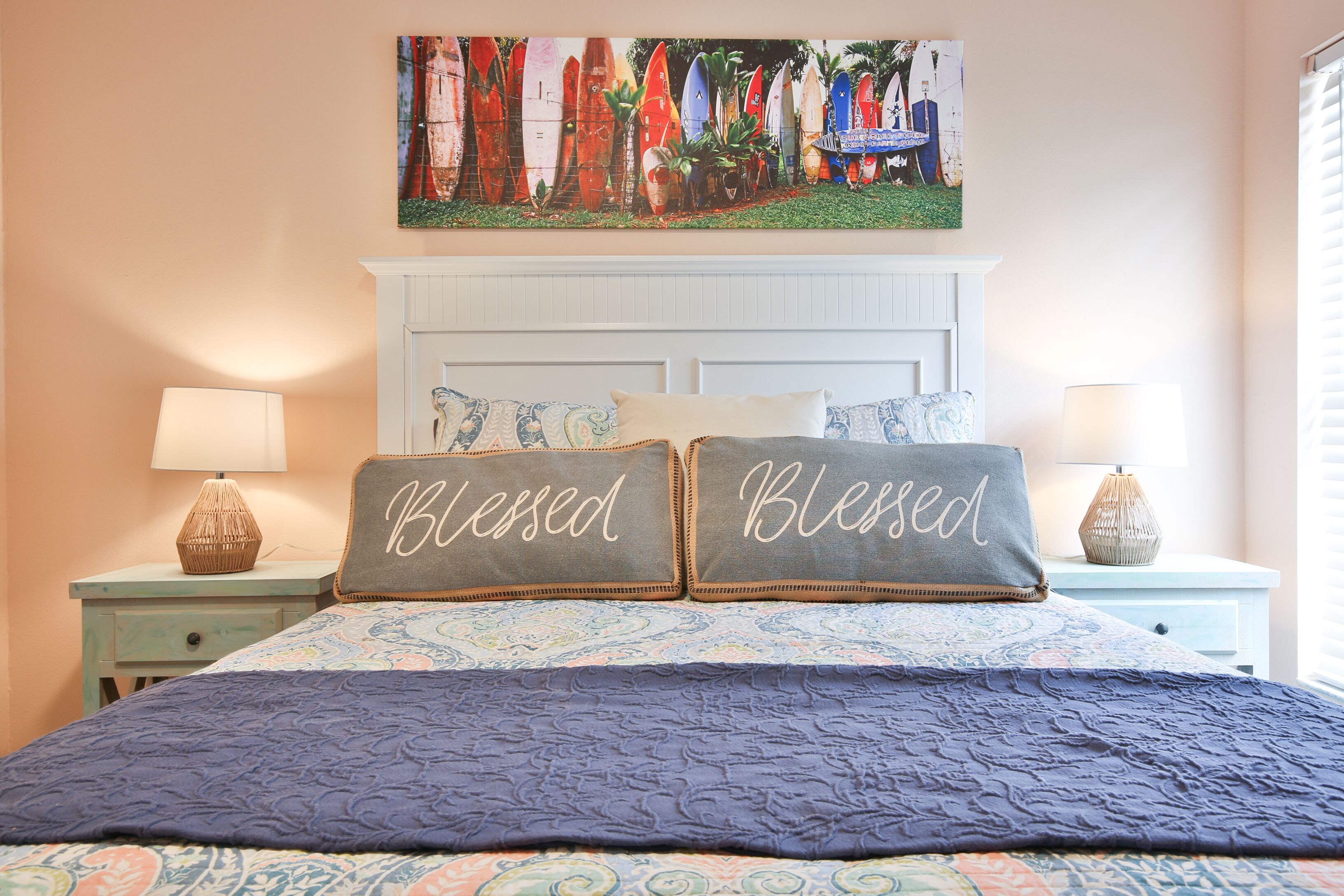 Une chambre à coucher avec un lit, deux oreillers ornés du mot « Béni », un tableau représentant des planches de surf accroché au mur, et une fenêtre avec des stores.