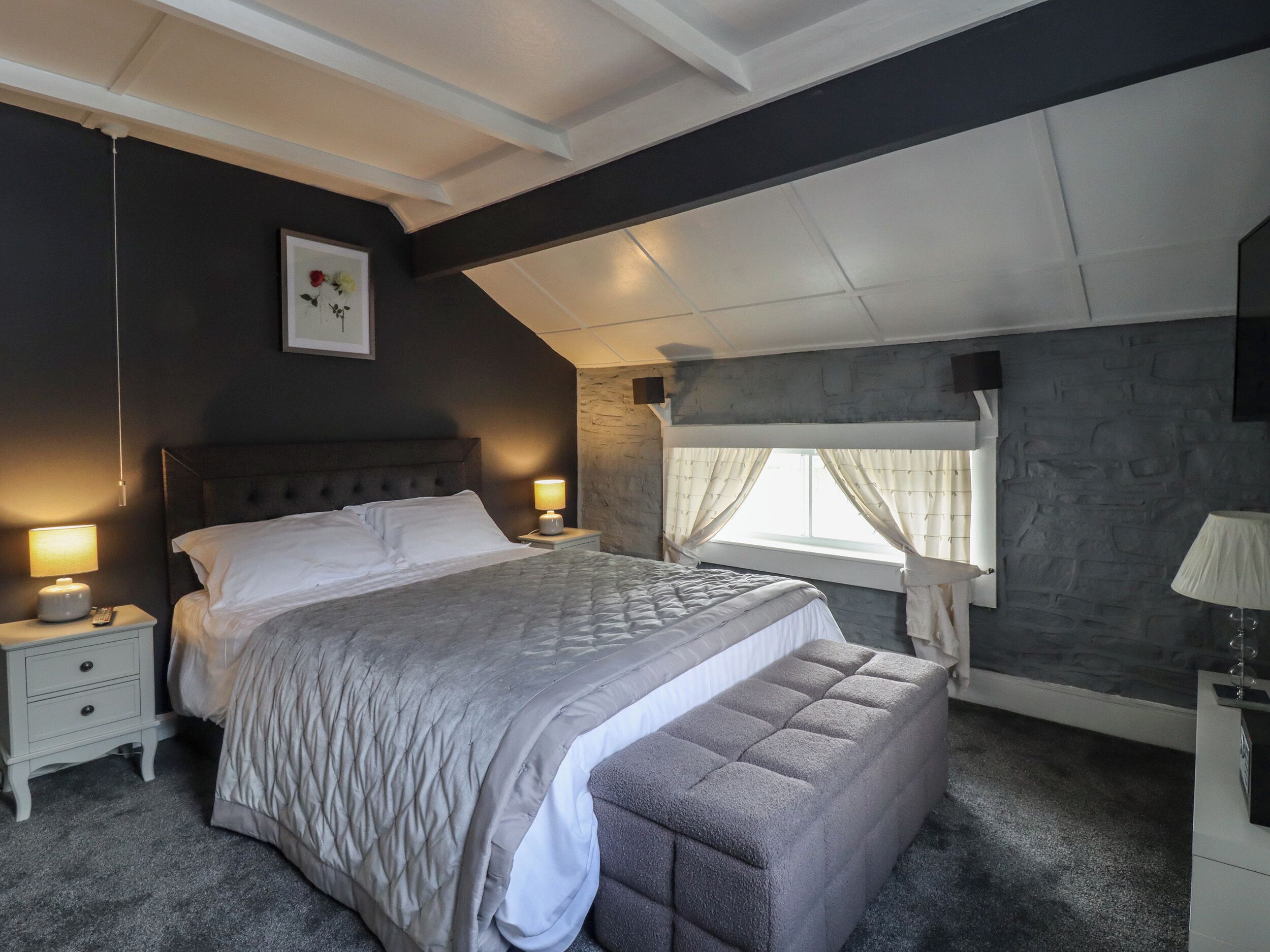 A bedroom with a large bed, bedside lamps, a nightstand, a window with curtains, and a stone wall.
