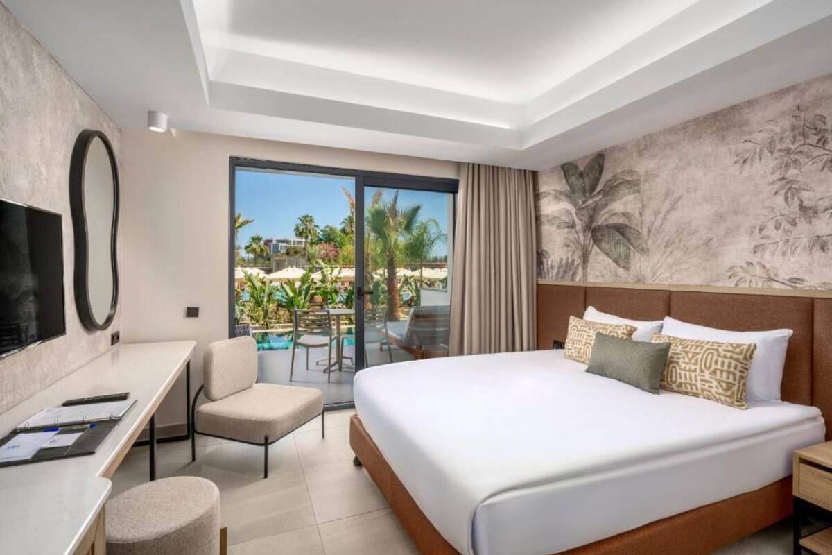 A modern hotel room with a large bed, a desk with a chair, a TV, and a balcony with a pool view.
