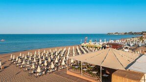 A beach with rows of sun loungers and umbrellas, a covered seating area, and a view of the sea and distant buildings. - Vox Maris Resort- Ultra All Inclusive (Manavgat)
