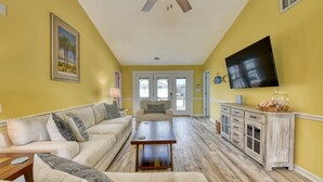 Un salon avec un ensemble de canapés de couleur beige, une table basse en bois, un téléviseur fixé au mur et un ventilateur de plafond.