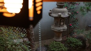 Un fanal de pedra en un jardí, amb una cadena, envoltat de plantes i una tanca de fusta.