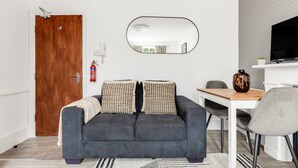 A modern living room with a grey sofa, a wooden door, a white wall, a mirror, a small table, and a television. - The Paddington Rooms (London)
