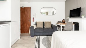 A modern hotel room with a kitchenette, a sofa with cushions, a dining area with a table and chairs, a flat-screen TV, and a large mirror. - The Paddington Rooms (London)