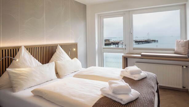 A modern hotel room with a large bed, a bench by the window, and a view of the sea. - Hotel Klippenklang (Helgoland)