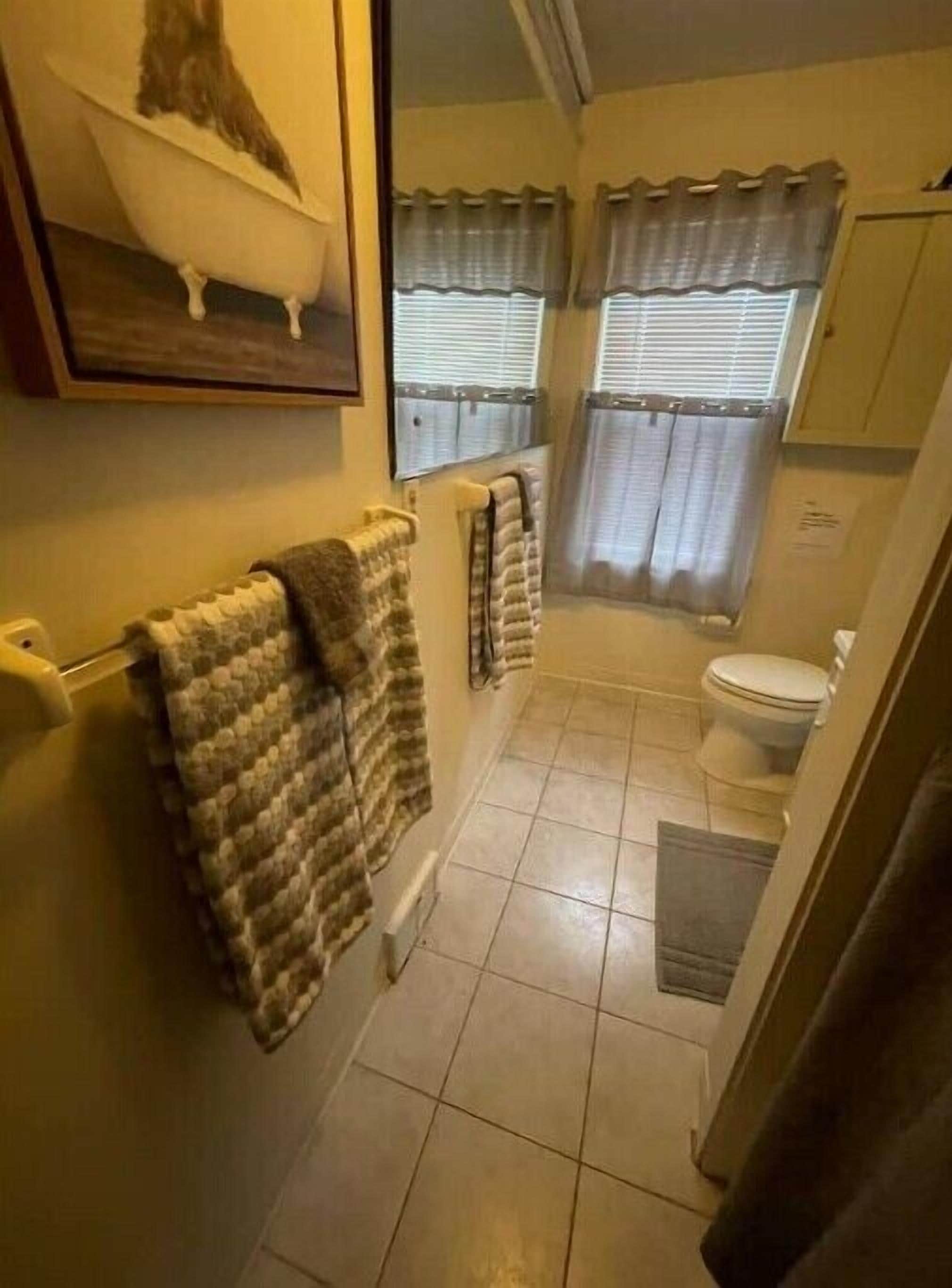 Une salle de bain équipée de toilettes, d’une baignoire et d’une douche. Elle comporte deux fenêtres avec des rideaux, et des serviettes sont disposées sur un porte-serviettes.