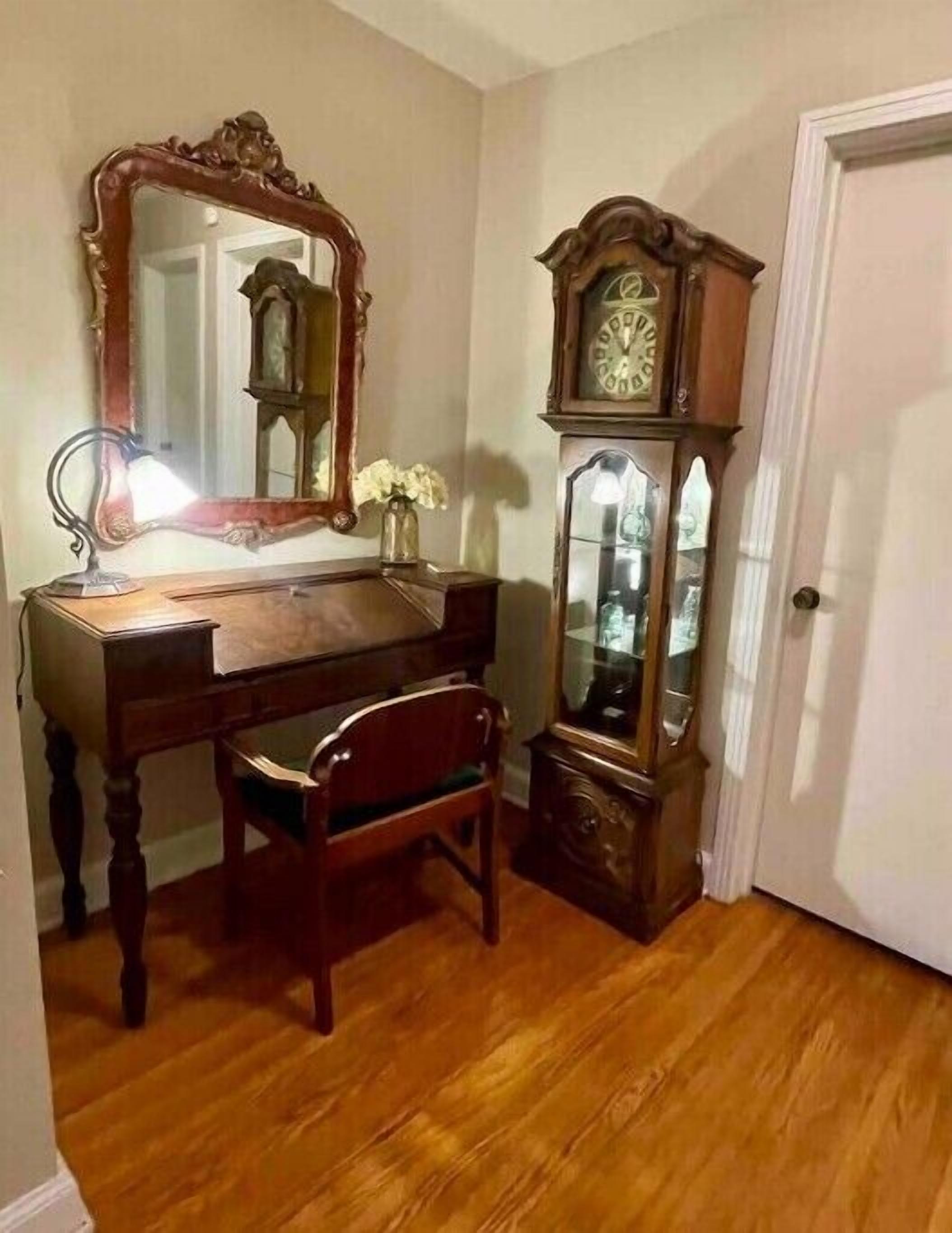 A wooden desk with a mirror and a clock, a chair, and a door in a room with wooden flooring.