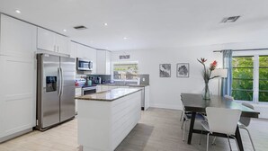 A modern kitchen with white cabinetry, a stainless steel refrigerator, and a dining area with a dark table and white chairs.