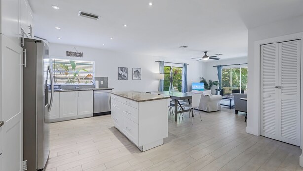A modern kitchen with white cabinetry and a central island, a dining area with a table and chairs, and a large window with curtains.