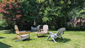 Ein Sitzbereich im Freien mit Adirondack-Stühlen und einem kleinen Tisch, umgeben von üppigem Grün und Bäumen.