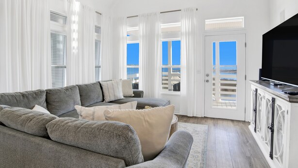 Un salon avec un canapé gris, des rideaux blancs, un téléviseur et une vue sur la mer par une porte.