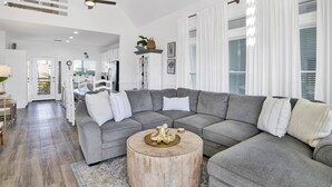 Un salon spacieux avec un grand canapé d’angle gris, une table basse ronde en bois et une grande fenêtre agrémentée de rideaux blancs.