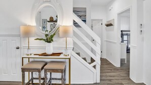 Un couloir avec un escalier blanc, une console surmontée d’un miroir, deux lampes et un banc.