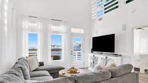 Un salon moderne avec un canapé d’angle gris, un téléviseur à écran plat et une vue sur la mer grâce à de grandes fenêtres.