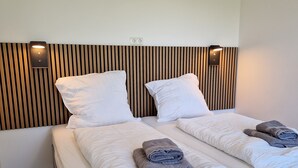 Quarto de hotel com duas camas, cabeceiras de madeira, roupa de cama branca e toalhas dobradas com capricho.