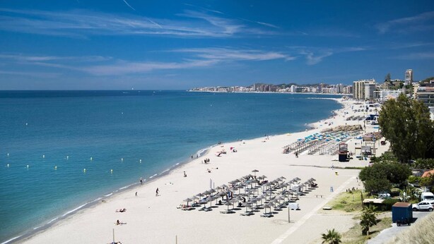 Ein Strand mit Reihen von Sonnenschirmen und Liegestühlen, einem klaren Himmel und einer Stadtlandschaft im Hintergrund.