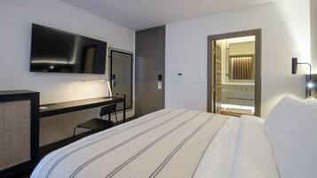 Una habitación de hotel con cama, escritorio, silla, televisor, puerta y baño visible a través de una puerta abierta.