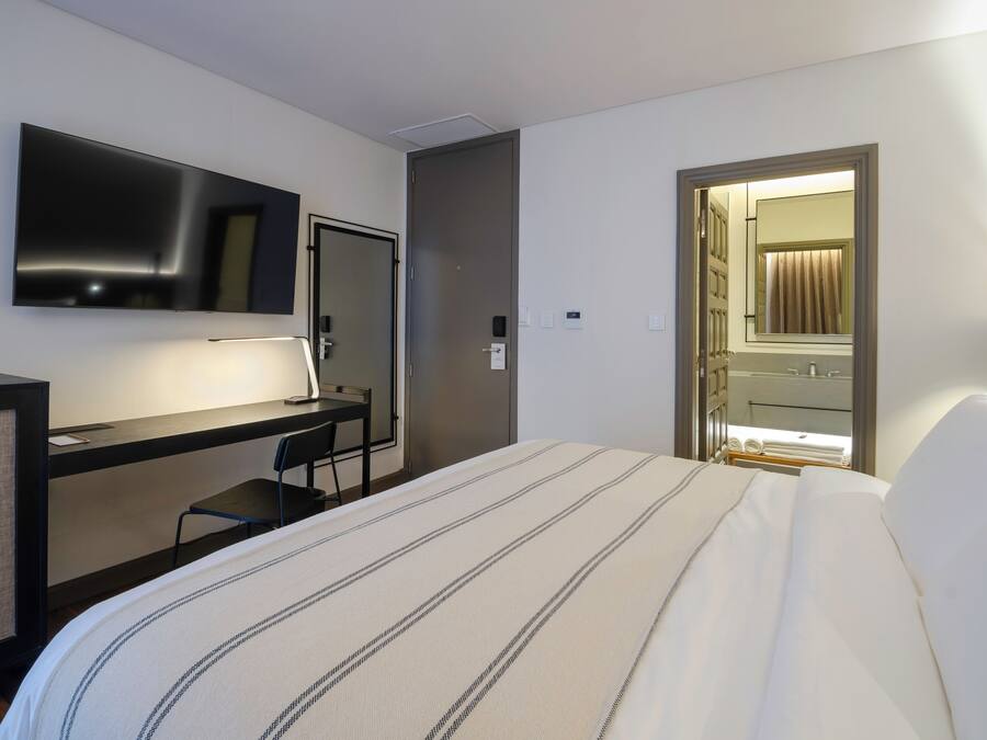 Una habitación de hotel con cama, escritorio, silla, televisor, puerta y baño visible a través de una puerta abierta.