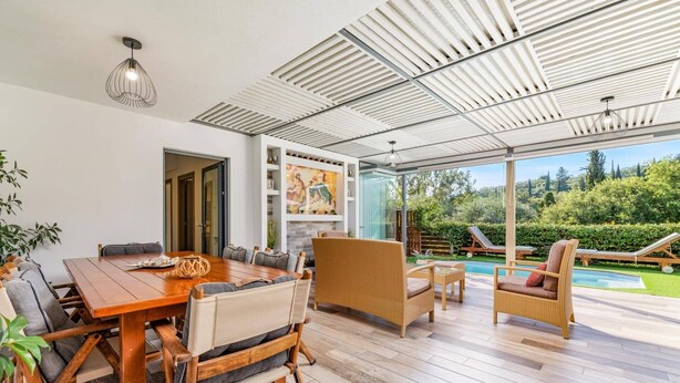 A modern outdoor dining area with a wooden table and wicker chairs, a glass-enclosed patio with a view of a swimming pool and greenery. - Villa Oras Corfu Dionisos by Villa Plus (Ionian Islands)