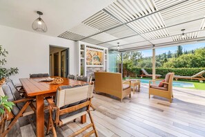Ett modernt utomhusområde för måltider med ett träbord och flätade stolar, samt en inglasad uteplats med utsikt över en swimmingpool och grönområden.