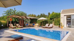 A rectangular swimming pool with lounge chairs and umbrellas, surrounded by greenery and a pergola with a lifebuoy.