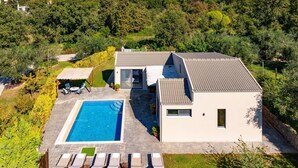Een modern huis met een zwembad, een terras met lounge stoelen en uitzicht op het omliggende bos en de bergen.