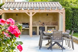 An outdoor kitchen with a tiled roof, a grill, and a dining area with chairs and a table set for two. - Villa Les Juliannes by Villa Plus (Azillanet)