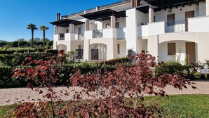 A multi-story building with balconies, a paved driveway, and a landscaped garden with red-leaved plants.