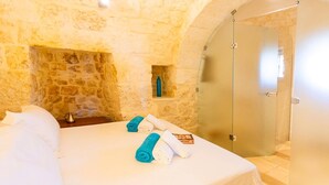 Ein Hotelzimmer mit Steinwand, einem Bett mit weißen Leinentüchern, zwei aufgerollten Handtüchern und einer Glastür zu einem Duschbereich.