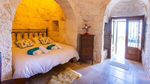 Une chambre à coucher avec un lit placé sous un plafond voûté en pierre, une commode en bois et une porte donnant sur l’extérieur.