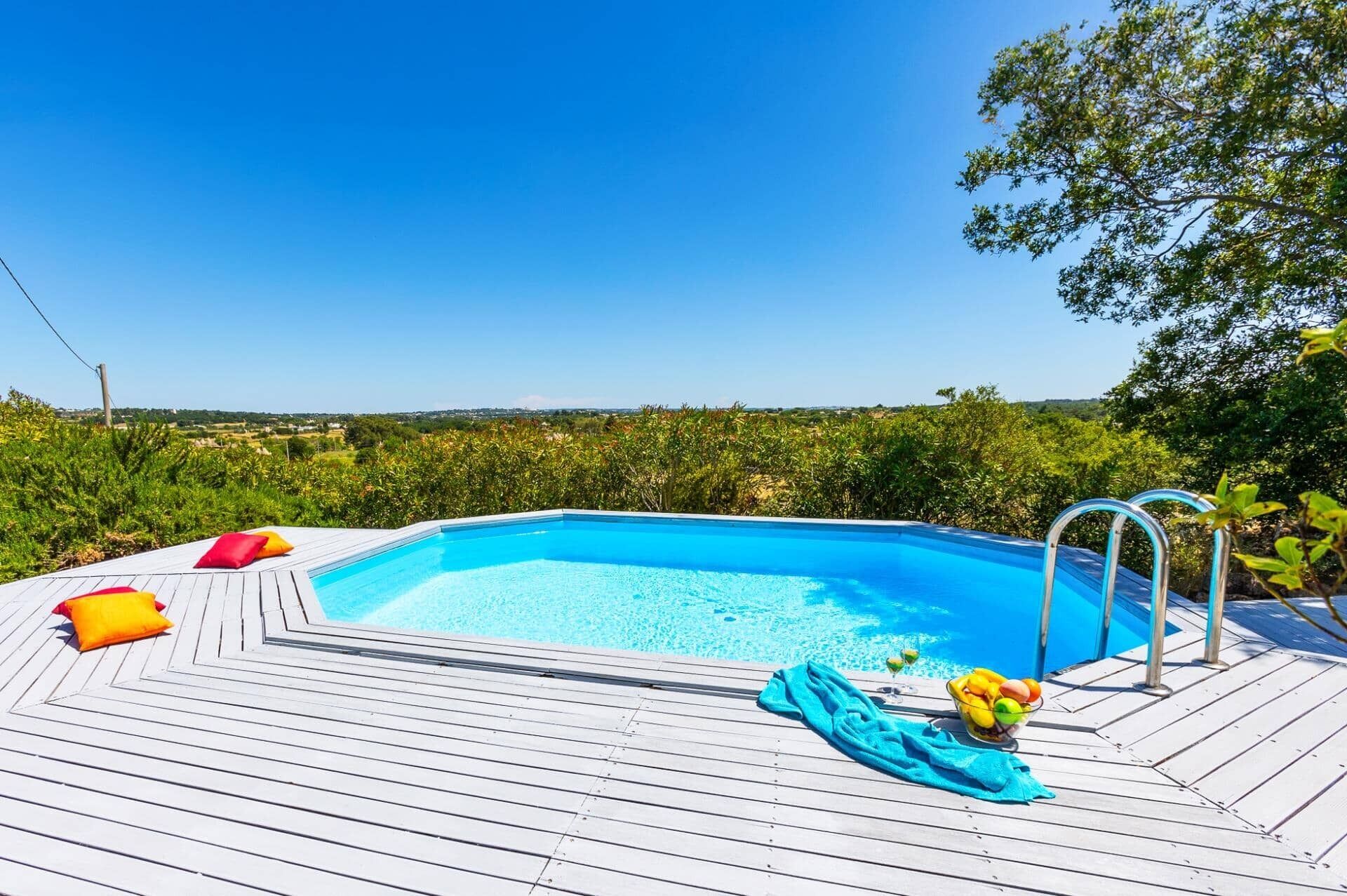 Une piscine dotée d’une terrasse en bois, entourée de verdure et surplombée par un ciel dégagé.