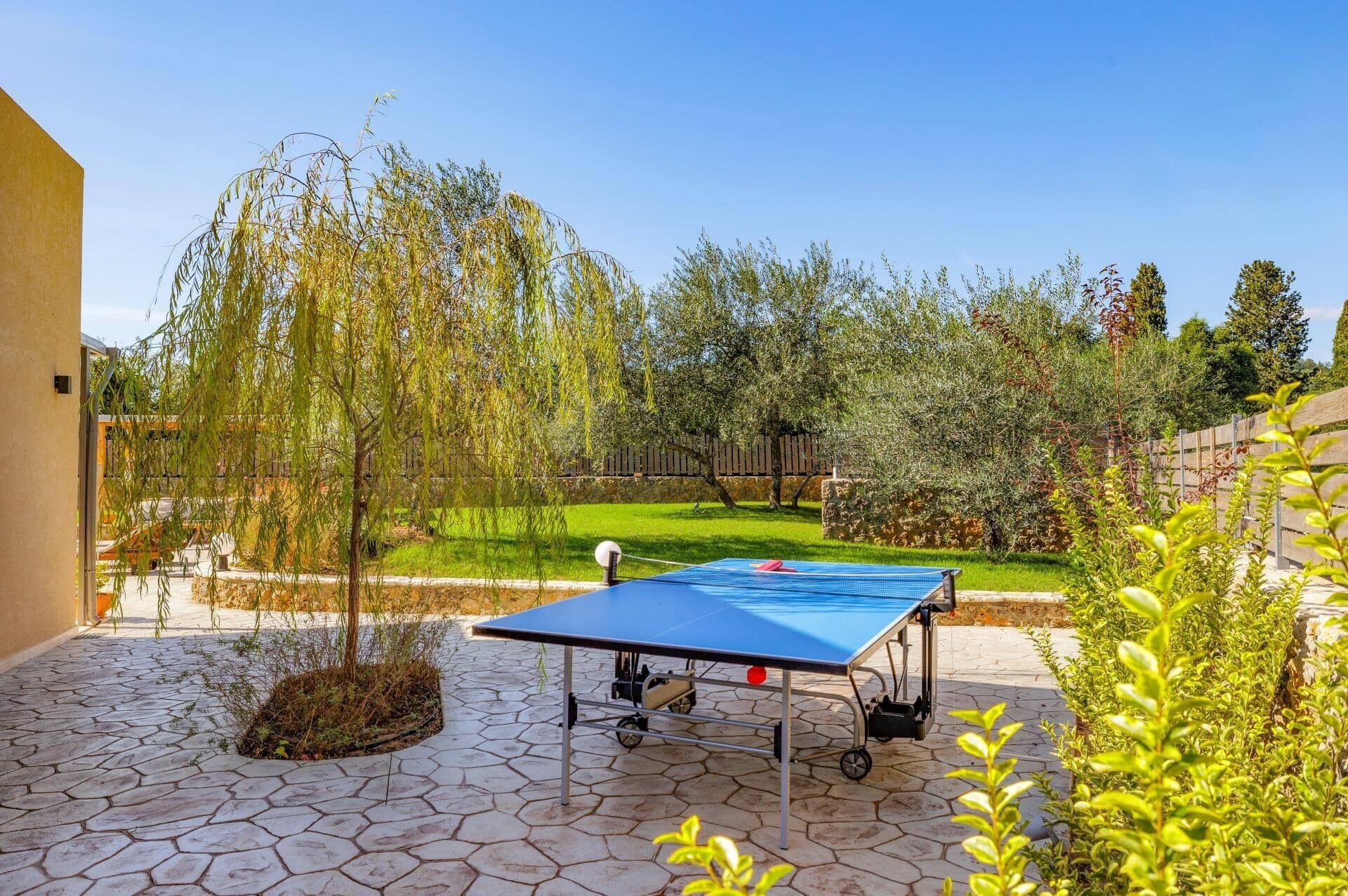 An outdoor ping pong table on a paved patio with a stone pattern, surrounded by greenery and trees.