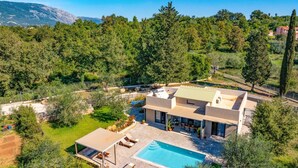 Eine moderne Villa mit Swimmingpool, umgeben von üppiger Vegetation und Bergen im Hintergrund.