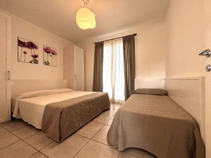 A hotel room with two beds, a large window with curtains, a wall-mounted light fixture, and framed floral artwork.
