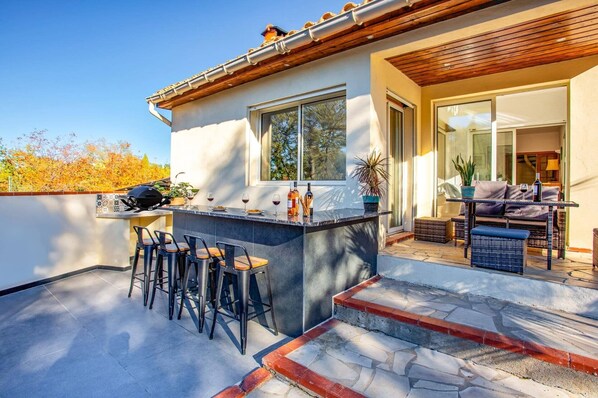 A modern outdoor patio with a bar area, seating, and a view of the surrounding trees. - Villa Des Alberes by Villa Plus (Laroque-des-Albères)